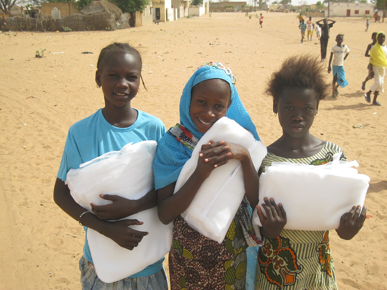 Three smiling girls standing in the sandy main square of a village in rural Senegal